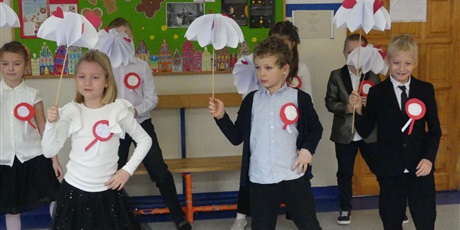 Powiększ grafikę: Grupa dzieci tańczy. Mają przyczepione biało-czerwone kotyliony, w dłoniach trzymają białe papierowe parasolki.