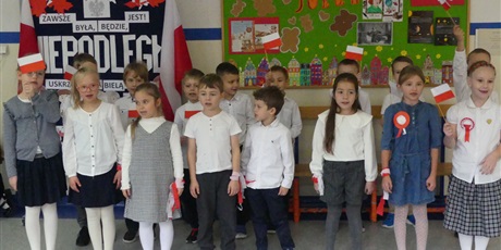 Powiększ grafikę: Grupa dzieci występuje na środku korytarza - śpiewają piosenkę. Część z nich trzyma w dłoniach małe biało-czerwone flagi.