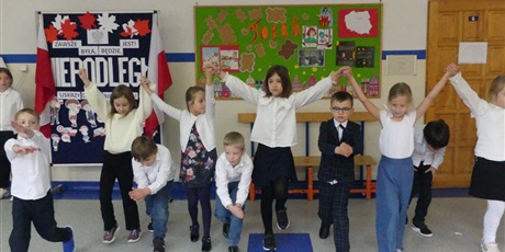 Powiększ grafikę: Grupa dzieci tańczy na środku korytarza. Część z nich trzyma się wysoko za dłonie - pod nimi przechodzą dzieci.