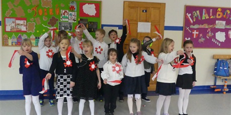 Powiększ grafikę: Grupa dzieci występuje na środku korytarza. Mają w dłoniach długie biało- czerwone bibułki - machają nimi.