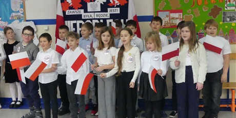 Powiększ grafikę: Grupa dzieci występuje na środku korytarza - śpiewają piosenkę, machają biało-czerwonymi flagami.