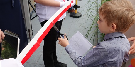 Powiększ grafikę: Chłopiec przecina nożyczkami biało-czerwoną wstęgę.