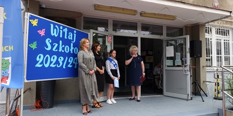 Powiększ grafikę: Przed budynkiem szkoły stoją trzy panie dyrektor i przewodnicząca Samorządu Uczniowskiego. Pani Dyrektor trzyma w dłoni mikrofon.