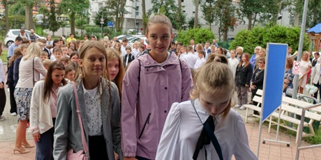 Powiększ grafikę: Grupa dzieci elegancko ubranych wchodzi do szkoły. Za nimi stoją rodzice.