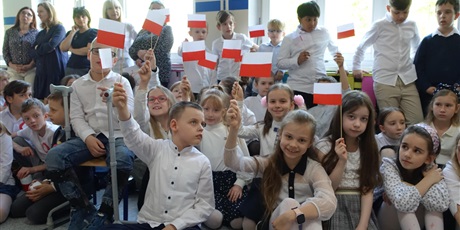 Powiększ grafikę: Grupa dzieci ubranych na galowo. W dłoniach trzymają biało-czerwone flagi.