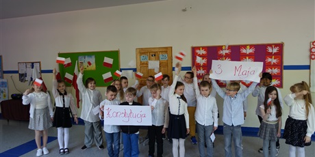 Powiększ grafikę: Grupa dzieci stoi na korytarzu, występuje przed innymi. W dłoniach mają flagi biało-czerwone i plakat z napisem Konstytucja 3 Maja.