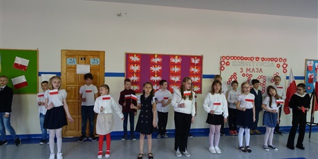 Powiększ grafikę: Grupa dzieci stoi na korytarzu, występuje przed innymi. W dłoniach mają flagi biało-czerwone.