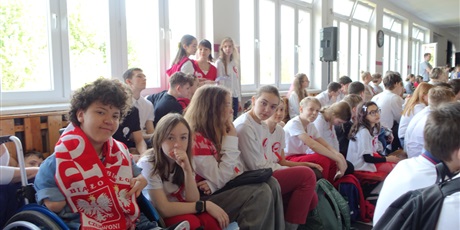 Powiększ grafikę: Grupa dzieci siedzi, są ubrani na biało-czerwono. Chłopiec ma ubrany szalik z napisem Polska.