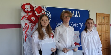 Powiększ grafikę: Chłopak i dwie dziewczyny stoją na środku korytarza, za nimi udekorowana tablica w biało-czerwone kwiaty.