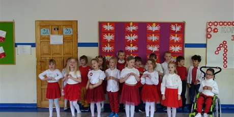 Powiększ grafikę: Grupa dzieci ubranych na biało-czerwono. Stoją na środku korytarza, występują.