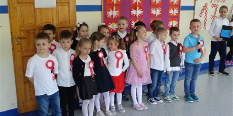 Powiększ grafikę: Grupa dzieci ubranych na galowo, z biało-czerwonymi kotylionami przypiętymi na piersi. Stoją na środku korytarza, występują.